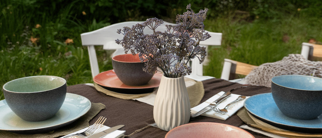 Ein stilvoll gedeckter Tisch im Freien mit farbigem Keramikgeschirr in Blau, Grün, Rosa und Braun, einer weißen Vase mit Trockenblumen, dunkler Tischdecke und weißem Besteck auf einer grünen Wiese.